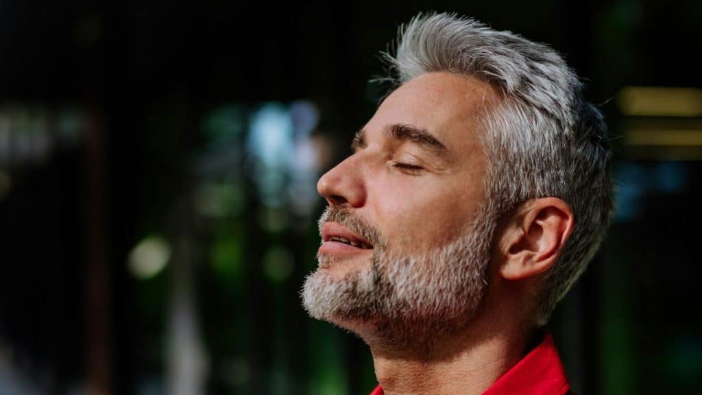A close-up side profile of a man with gray hair and a beard, eyes closed, face tilted up, wearing a red shirt in shadow.