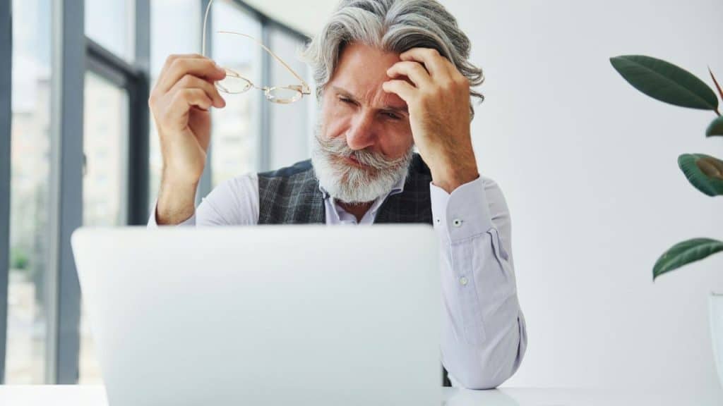 Older man with a white beard holds glasses while frowning at a laptop screen.