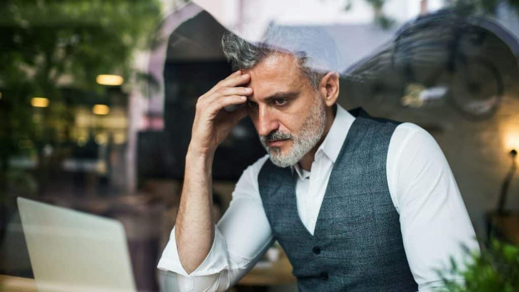 Man with a beard and vest holds his forehead while looking at a laptop through a window.