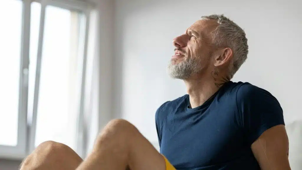 Older man with gray beard looking up, sitting with bent knees by a bright window.