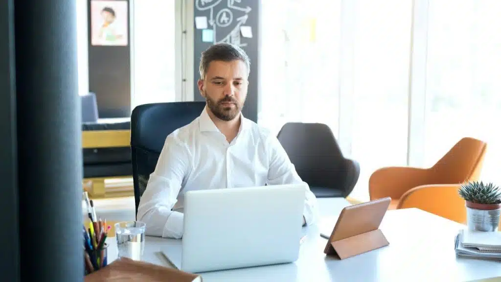 A focused man with a short beard and white shirt sits at a modern office desk, looking down at his laptop.