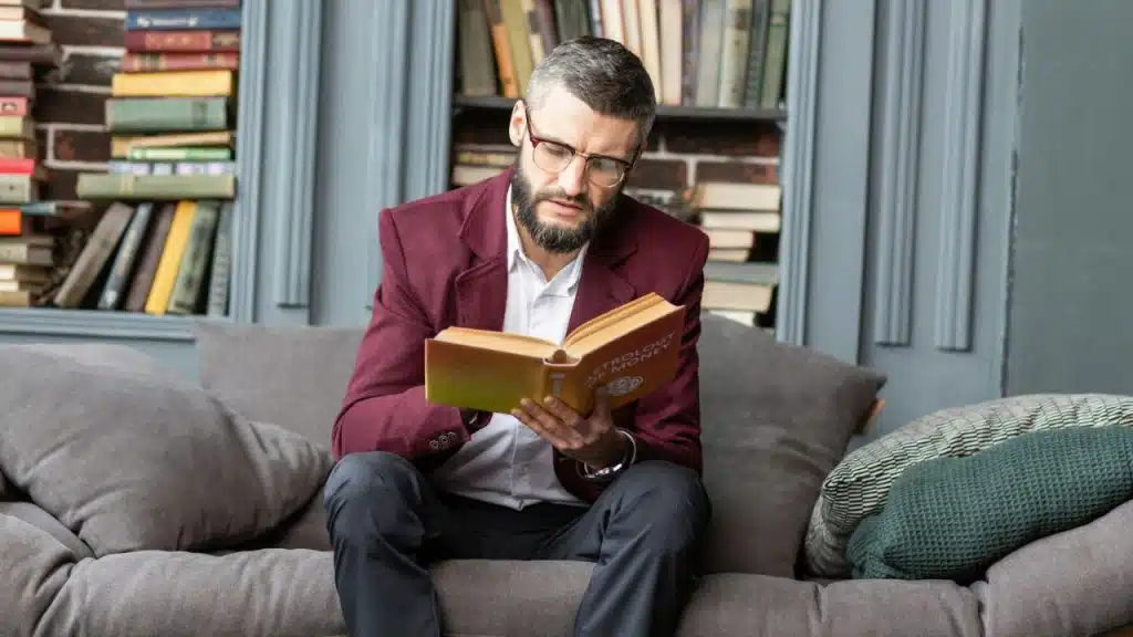 Man in a burgundy jacket and glasses sitting on a couch reading a book.