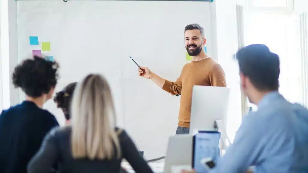 Smiling man with a beard pointing at a whiteboard while presenting to a group of people.