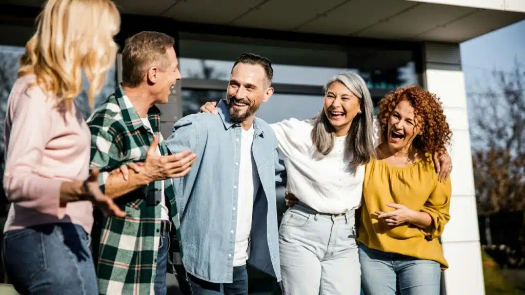 Five middle-aged adults laughing and hugging outside a modern building on a sunny day.
