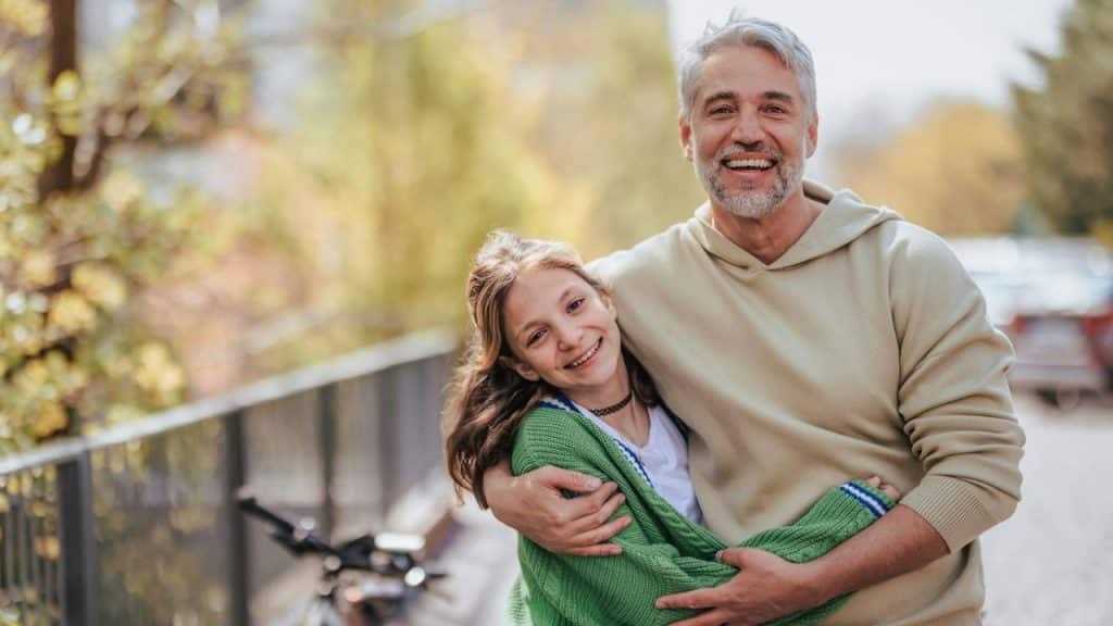 Smiling man with gray hair hugging a younger girl in a green sweater outdoors.