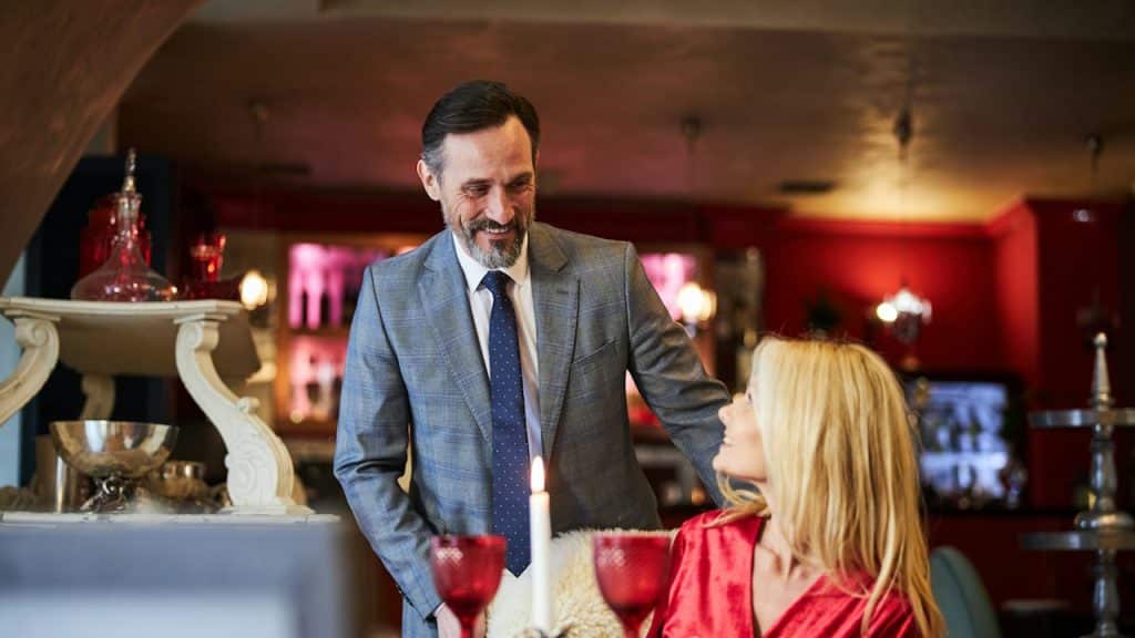 A formally dressed man standing over a table, smiling and looking down at a blonde woman in a red outfit, with a lit candle and wine glasses on the table between them.