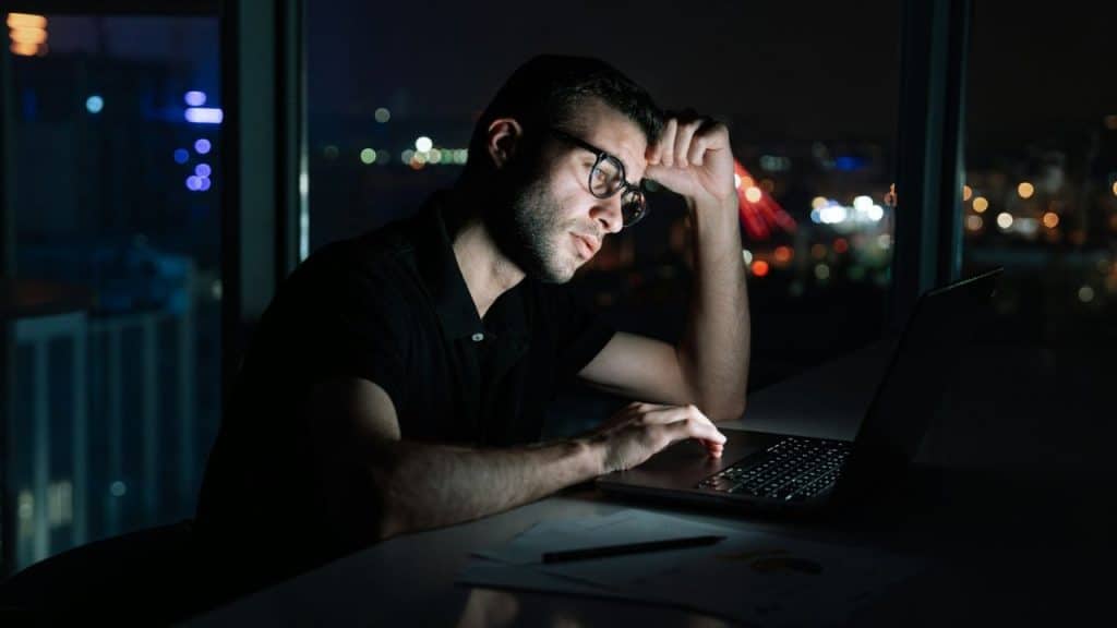 A man wearing glasses is working at a laptop late at night with a brightly lit city skyline in the background.