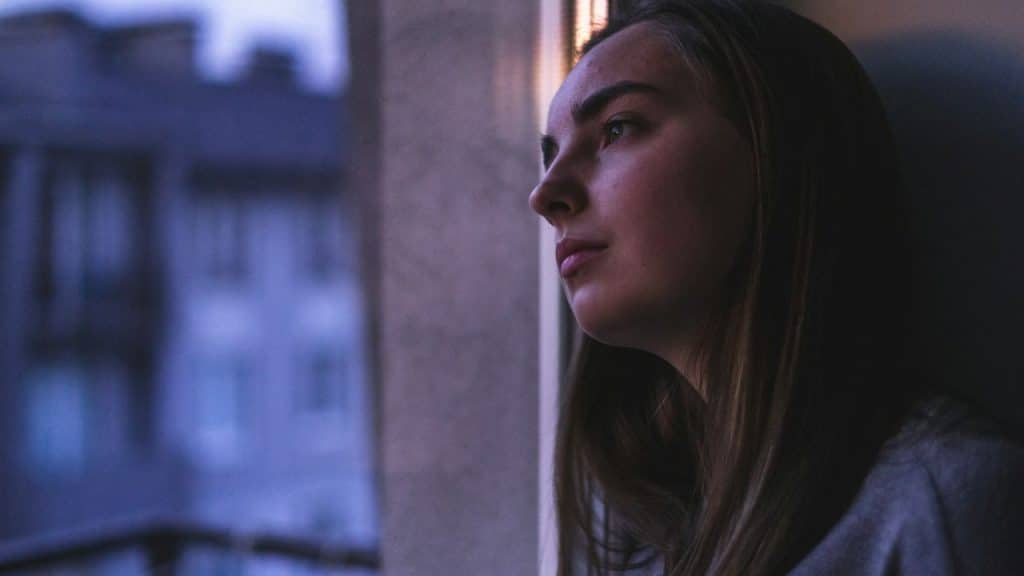 A young woman looks out a window in dim light, with a blurred cityscape visible outside.