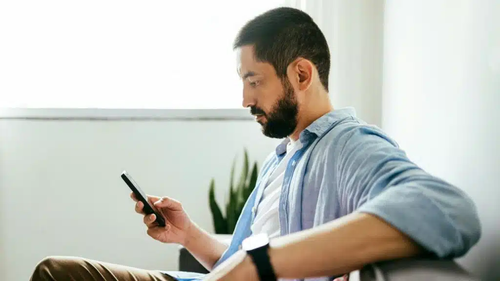 A bearded man in a light blue shirt sits in a chair and looks at his smartphone.