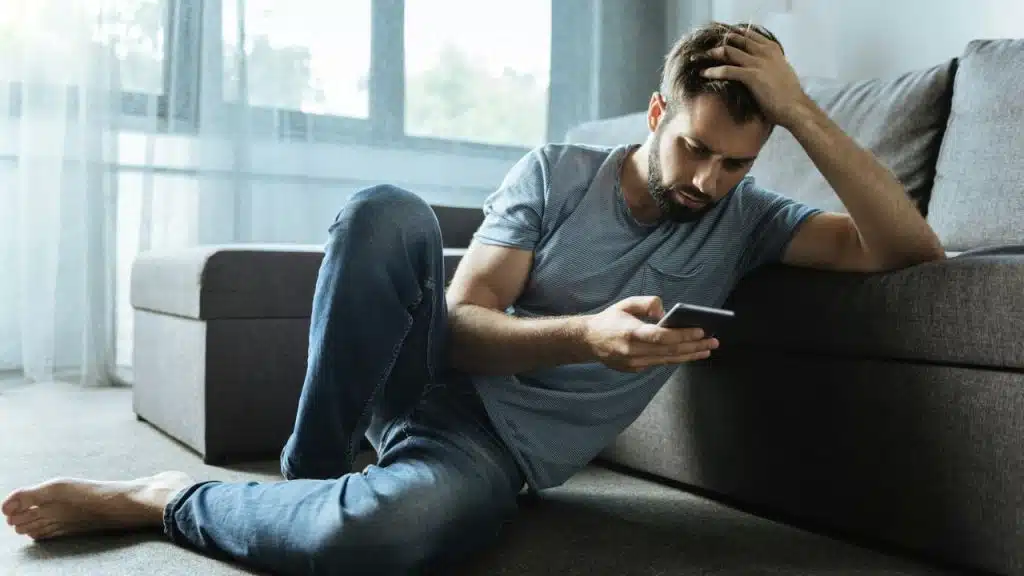 A man sits on the floor by a sofa, looking distressed while checking his phone.