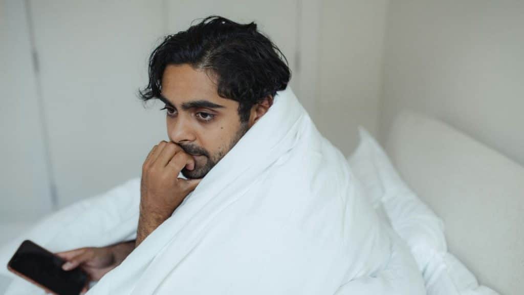 Man sitting up in bed, wrapped in a white blanket, holding a phone.