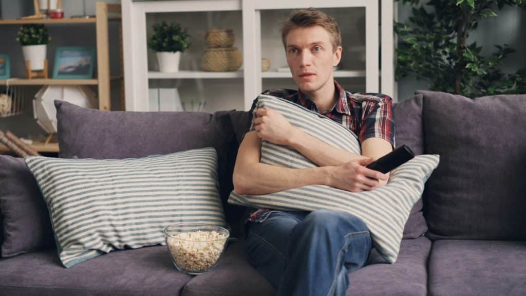 A young man is sitting on a sofa, hugging a pillow and holding a remote, with a bowl of popcorn nearby.