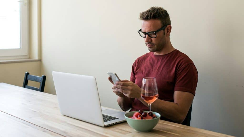 A man in a red shirt and black glasses looks at a phone while sitting at a table with a laptop, wine glass, and bowl of fruit.