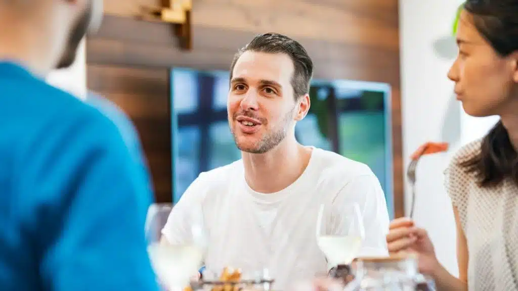 A smiling man in a white t-shirt is talking to people around a table, holding a glass of white wine.