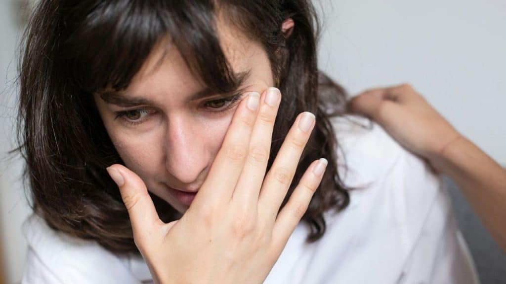 Close-up of a distressed woman with dark hair, covering one eye with her hand.