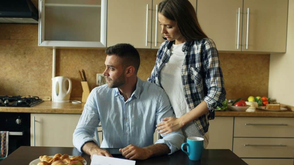 A woman in a plaid shirt stands behind a man seated at a table, resting her hand on his shoulder in a kitchen setting.