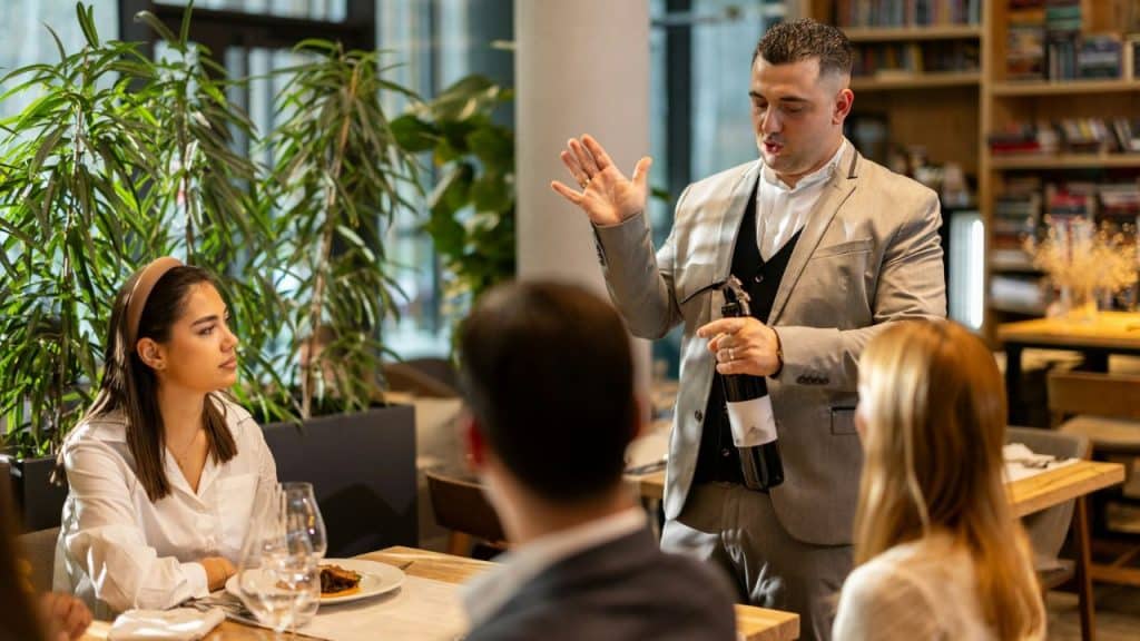 A man in a light suit holds a wine bottle and talks to people seated around a restaurant table.