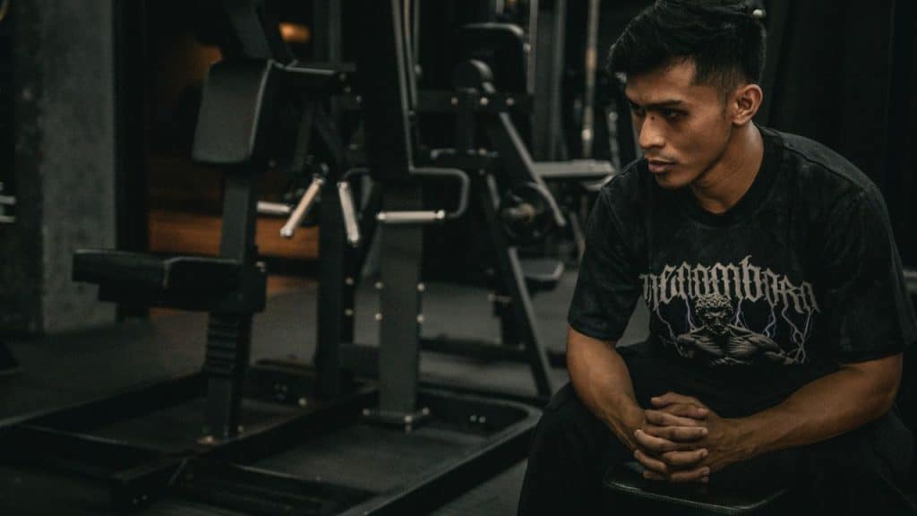 Young man in a black graphic t-shirt sitting on a bench in a dimly lit gym.