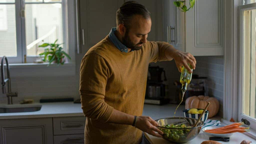 Bearded man pouring olive oil on vegetables in a bowl in a bright kitchen.