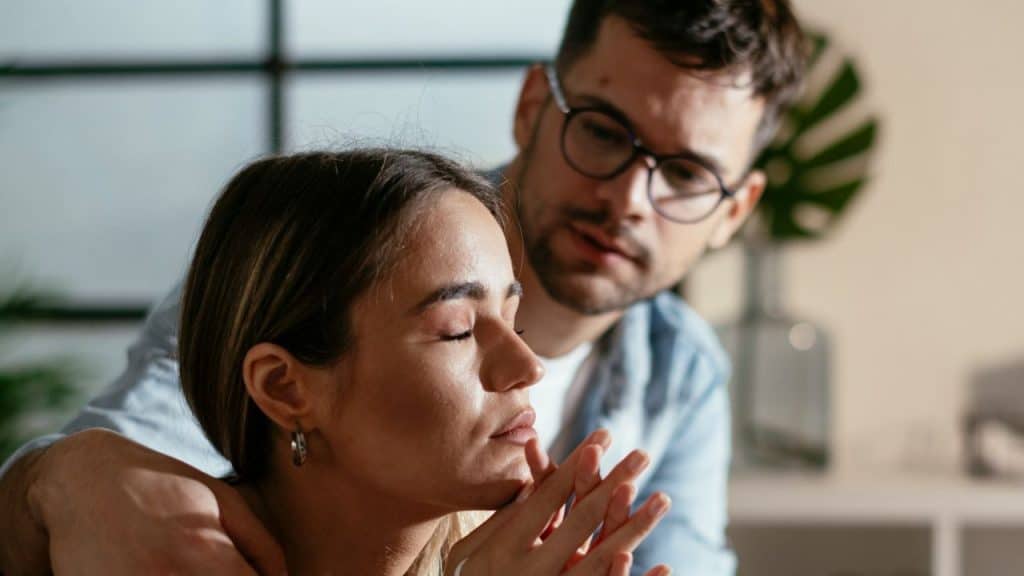 Man with glasses standing over a woman with her eyes closed, his hand on her shoulder.