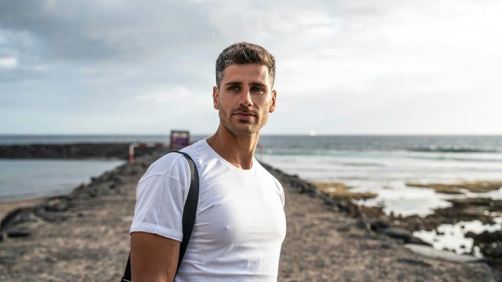 A man in a white t-shirt standing by the ocean.