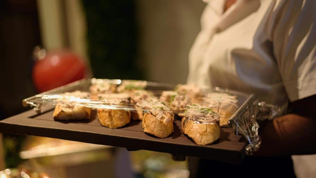A server holding a tray of small baguette appetizers covered with plastic wrap.
