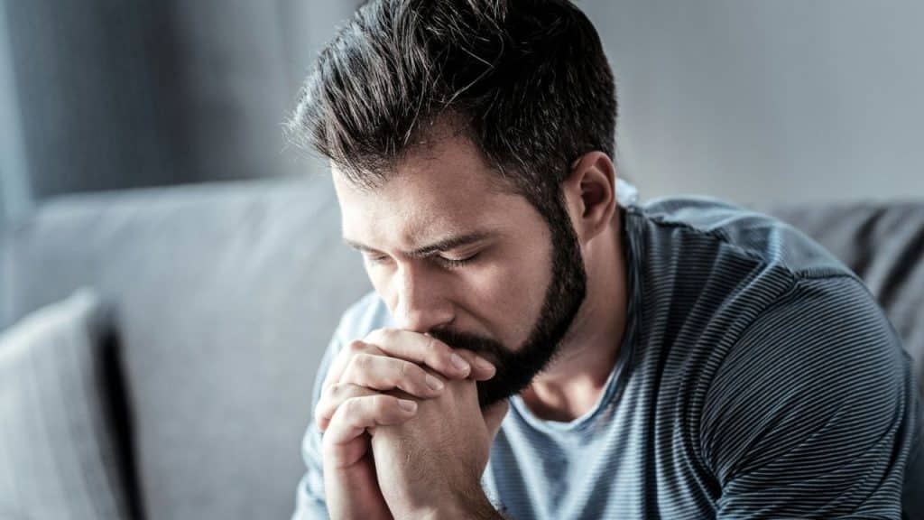 A man with dark hair and a beard sits indoors, his eyes closed, with his hands clasped under his chin in a pensive posture.