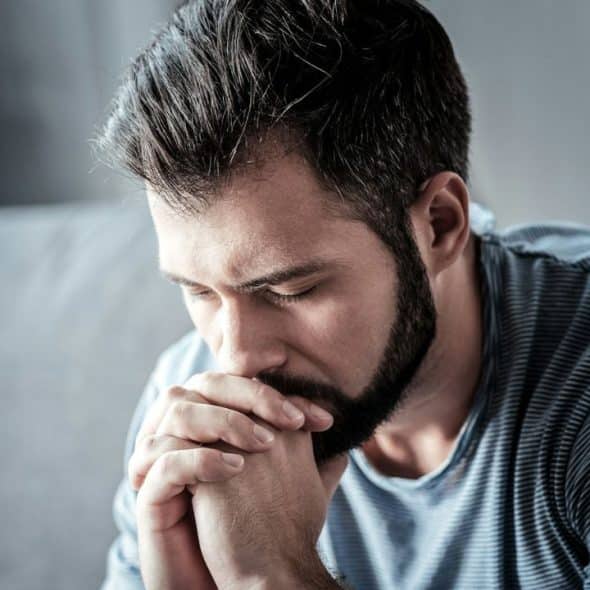 A man with dark hair and a beard sits indoors, his eyes closed, with his hands clasped under his chin in a pensive posture.