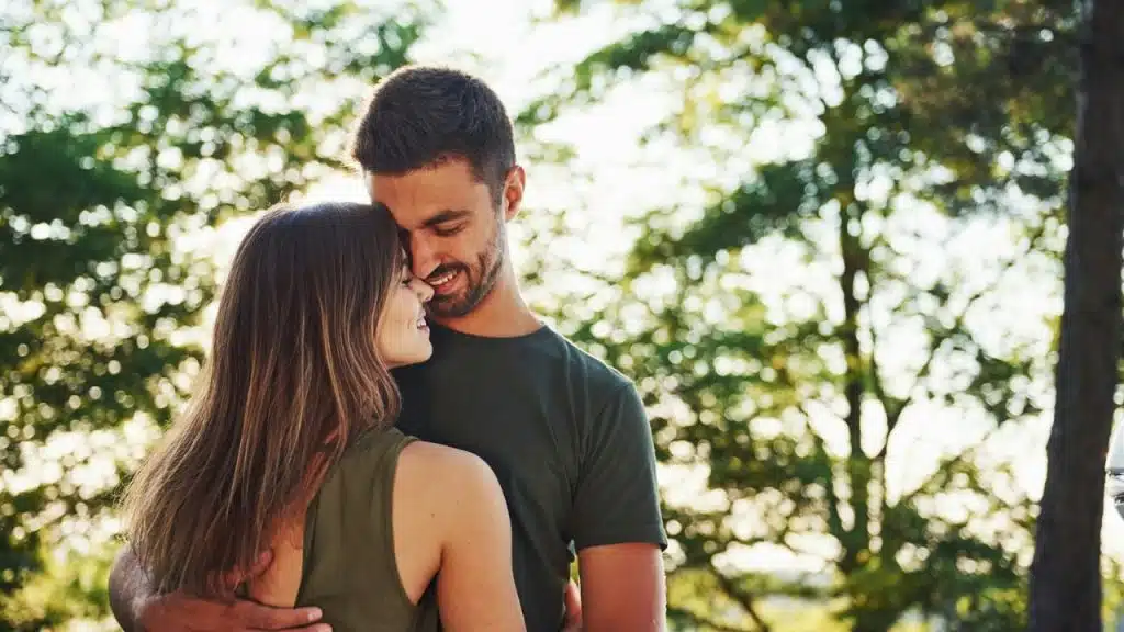 A man and a woman embracing outdoors in front of green, sunlit trees.