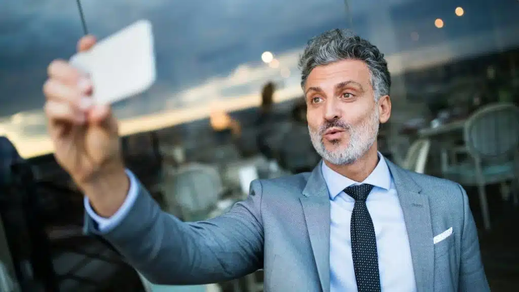 A stylish, gray-haired man in a suit and tie takes a selfie outdoors.