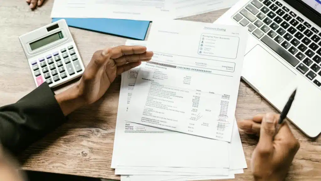 A person reviewing financial documents with a calculator and a laptop on the table.