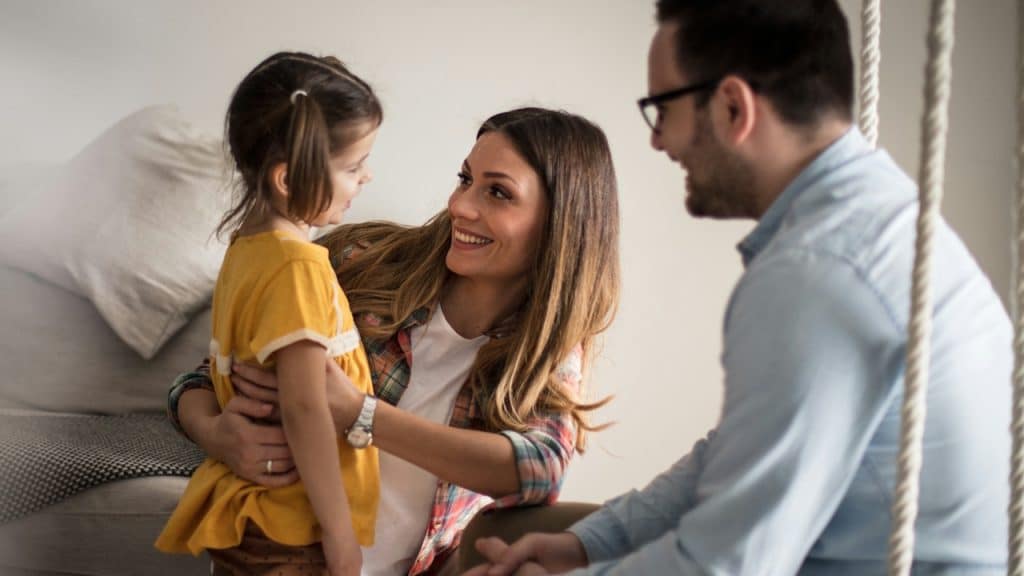 A young couple, smiling, sits on a couch while the mother holds a young girl with pigtails in a yellow shirt.