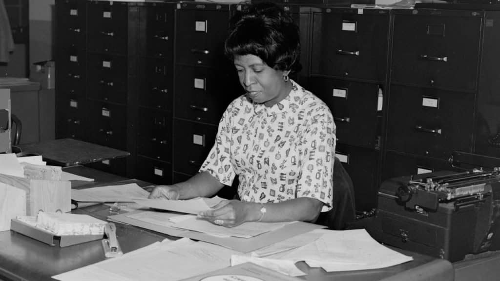 A black and white photo of a woman with short dark hair seated at a desk covered in papers and a typewriter, with tall filing cabinets behind her.