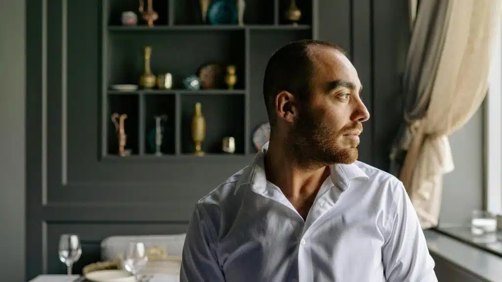 A man in a white shirt looking out of a window thoughtfully.