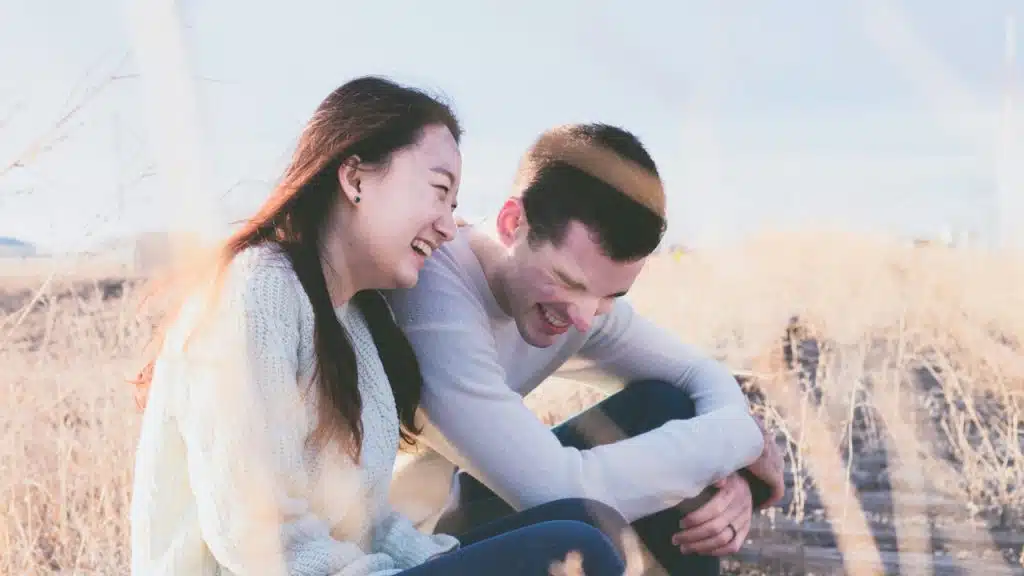 A happy couple sitting close together in a sunny field of dry grass, both laughing joyfully.