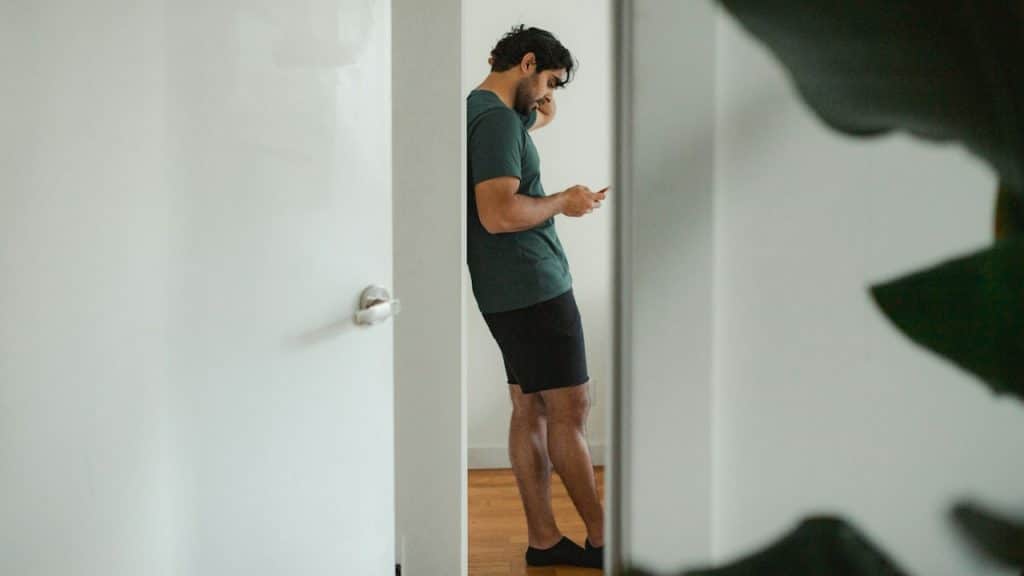 A man in a dark green t-shirt and black shorts stands in a doorway, leaning against the wall, looking down at his smartphone.