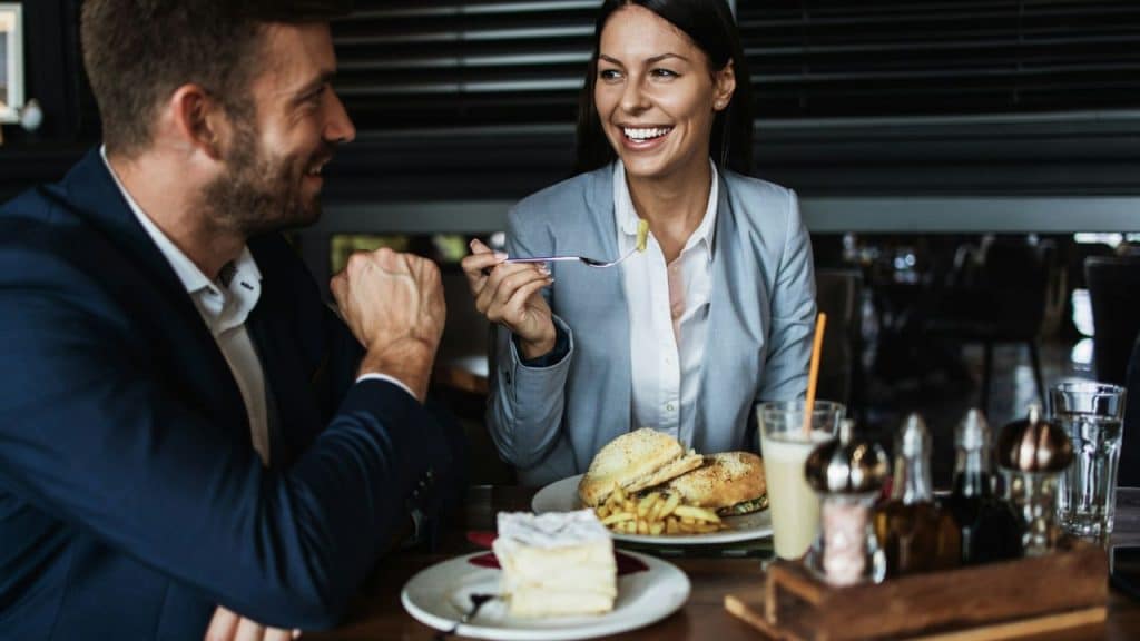 Man and woman in business attire smiling at each other while eating a meal.