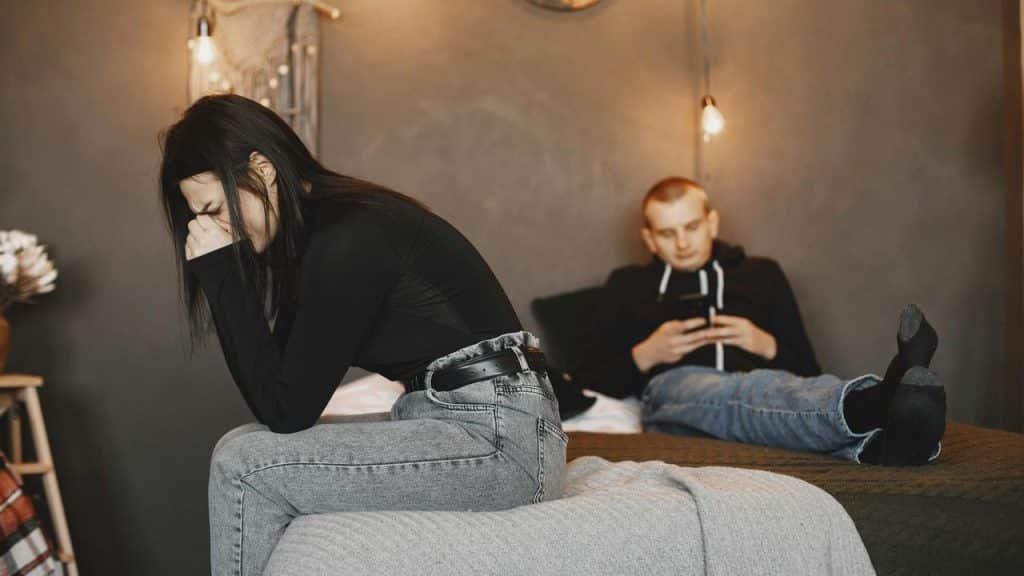 A woman sitting on a bed looking upset while a man uses his phone behind her.