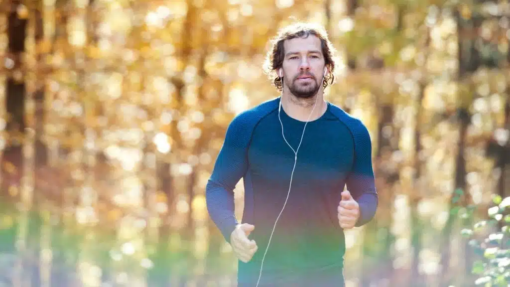 A man with earbuds is jogging outdoors in a forest with yellow and gold foliage.