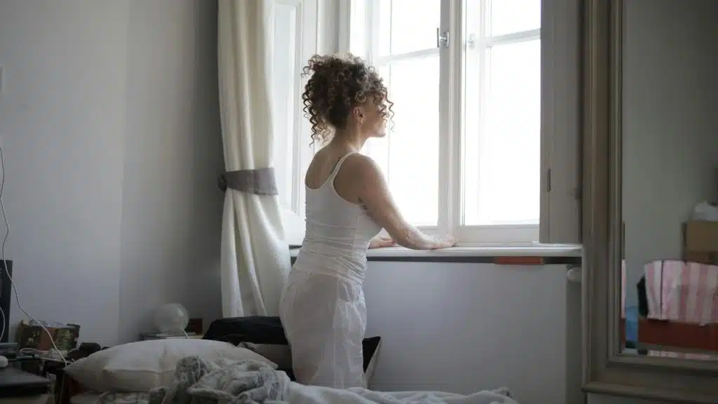A person with curly hair standing by a bright window in a bedroom.