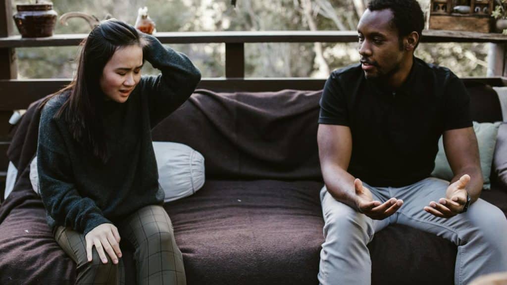 A couple sitting outside on a couch, with the woman upset and the man talking to her.