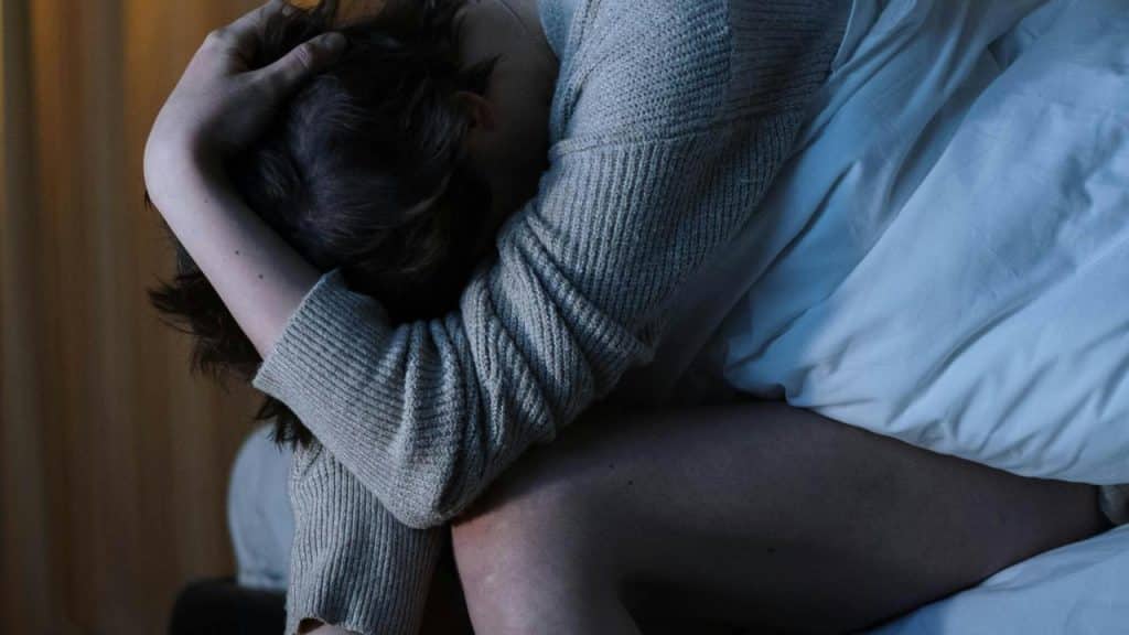 A person sitting on a bed with their head in their arms, appearing distressed.