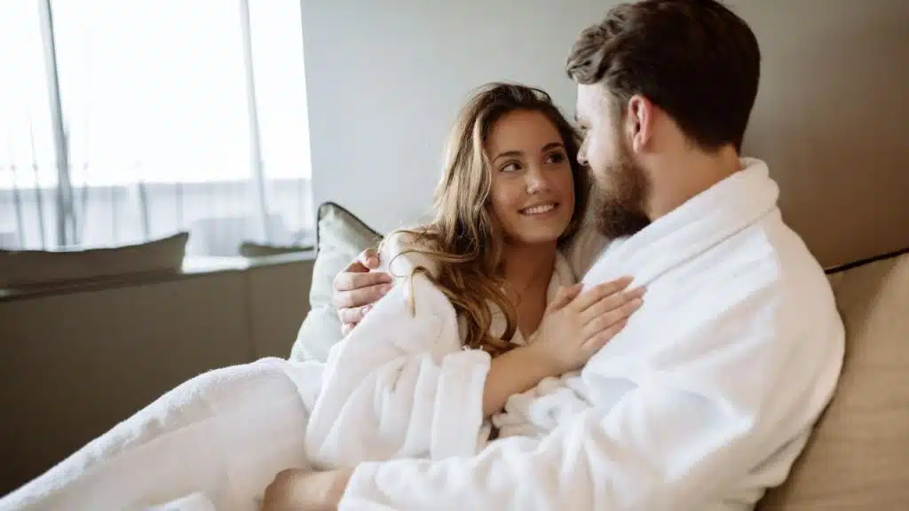 A man and woman in white bathrobes are sitting together and smiling at each other.
