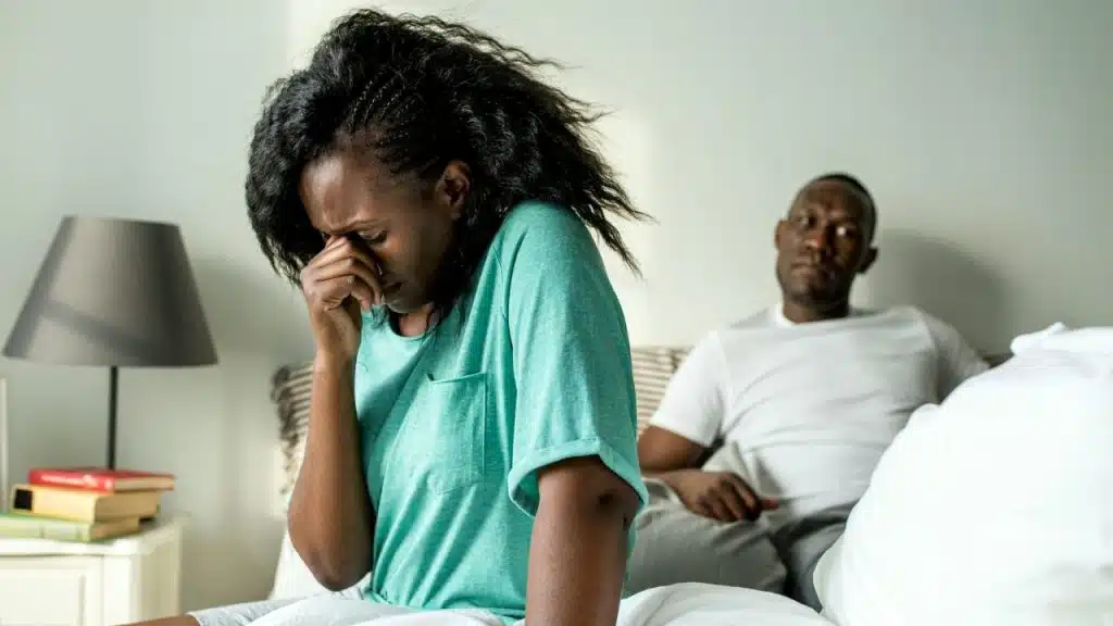 A distressed woman in a teal shirt sits on a bed, holding her hand to her face, while a man sits blurred in the background.