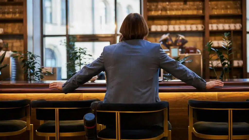 A man in a gray suit is sitting at a wooden bar, facing away from the camera.