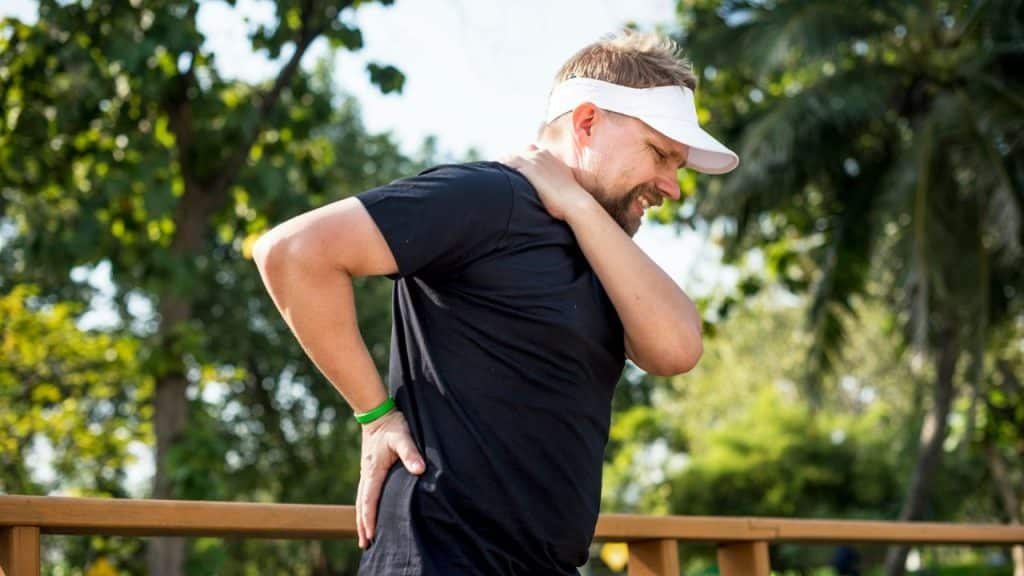 A man wearing a visor and black shirt is holding his back and neck in pain outdoors.