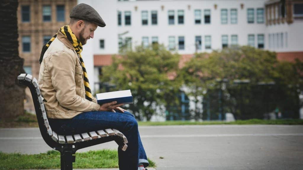 Man in a flat cap and scarf sitting on a park bench reading a book.