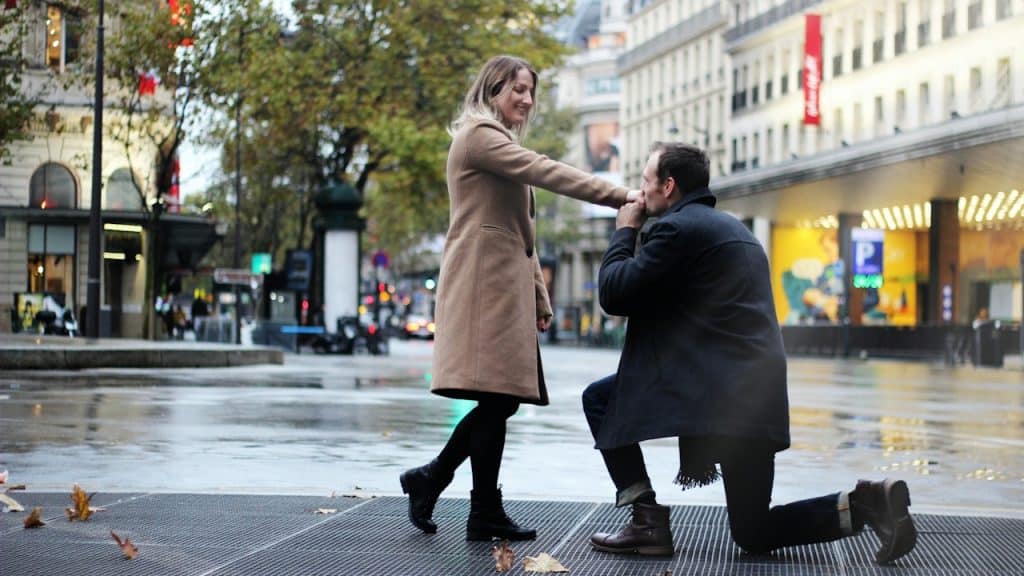 A man kneels on one knee on a wet city street to kiss the hand of a smiling woman in a tan coat.