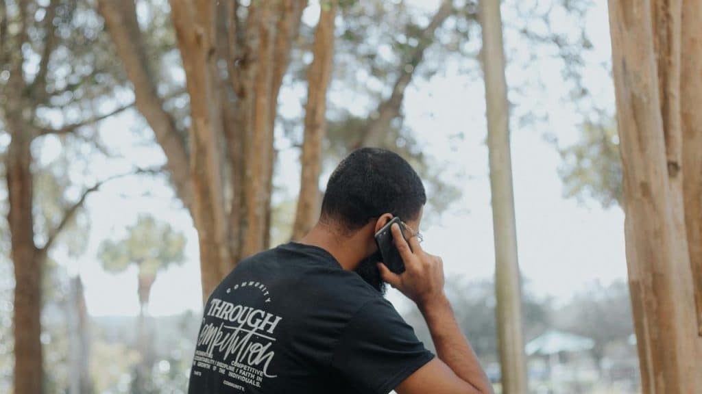 A man talking on his smartphone while standing outdoors among trees.