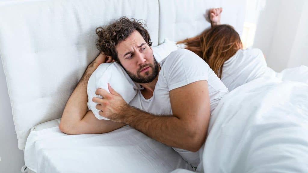 A worried man lies awake in bed, hugging a pillow while a woman sleeps facing away from him.